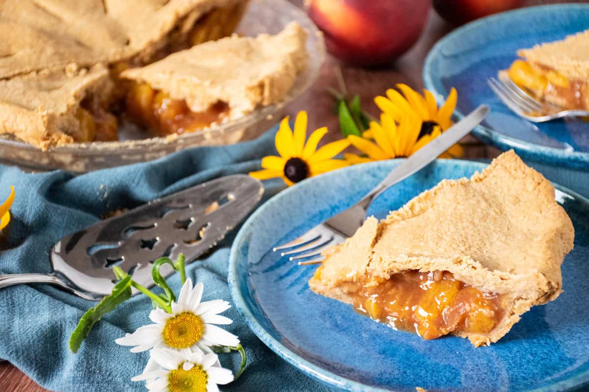 Slice of peach pie on a dessert plate, with the rest of the pie visible in the background.