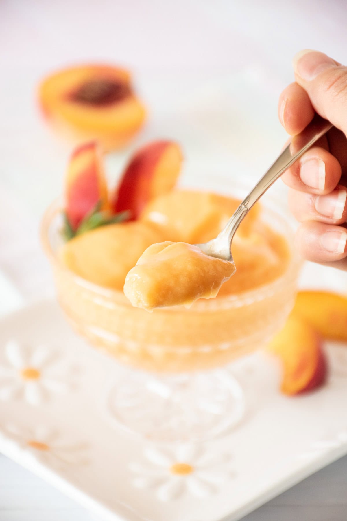 Glass dish with three scoops of peach sorbet; garnished with peach slices and fresh tarragon. A spoonful of sorbet is being held up in the foreground.