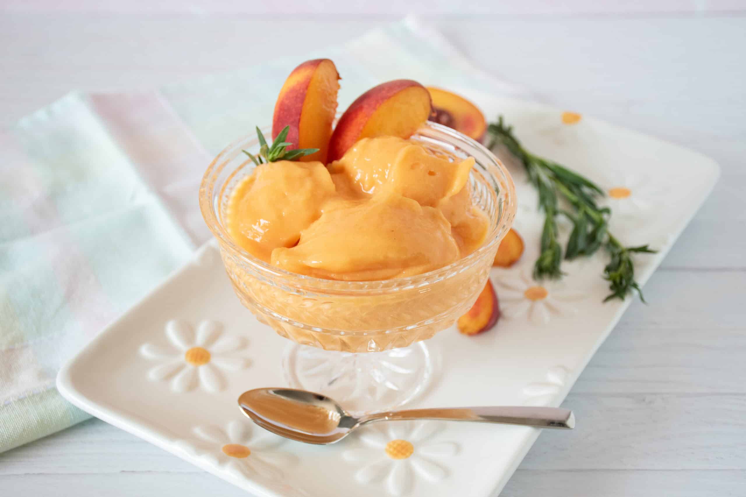Glass dish with three scoops of peach sorbet; garnished with peach slices and fresh tarragon. More fresh peach slices and sprigs of tarragon visible in the background.