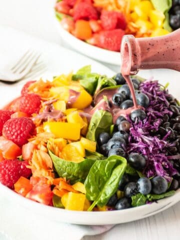 Salad with rainbow colored vegetables, and dressing being poured on.