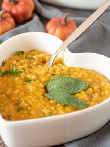 Pumpkin steel cut oats risotto in a heart shaped bowl, garnished with fresh sage leaves and chopped parsley.