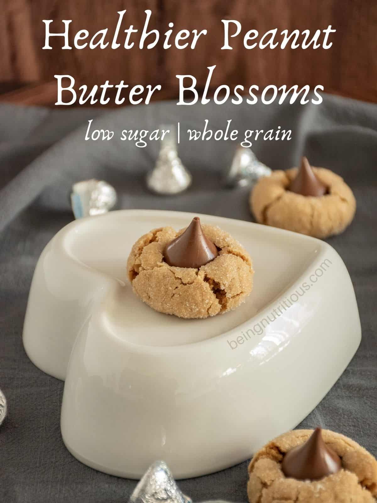 Peanut butter blossom on a heart shaped dish, with more cookies and kisses scattered around the table. Text overlay: Healthier peanut butter blossoms; low sugar, whole grain.