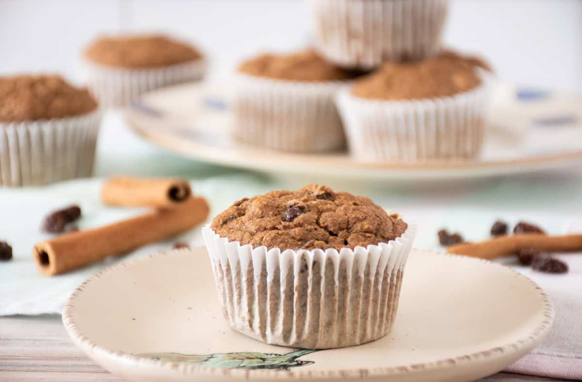 Single muffin on a dish. Another dish with a stack of muffins in the background. Cinnamon sticks and raisins are scattered around the table.