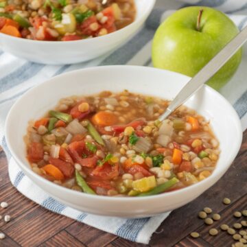 Bowl of soup with a spoon in it, and a green apple on the table behind it.