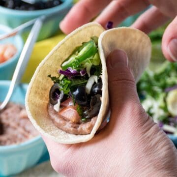 Close up image of 2 hands holding a taco, filled with refried beans, tomatoes, black olives, crunchy greens, and a jalapeno slice.