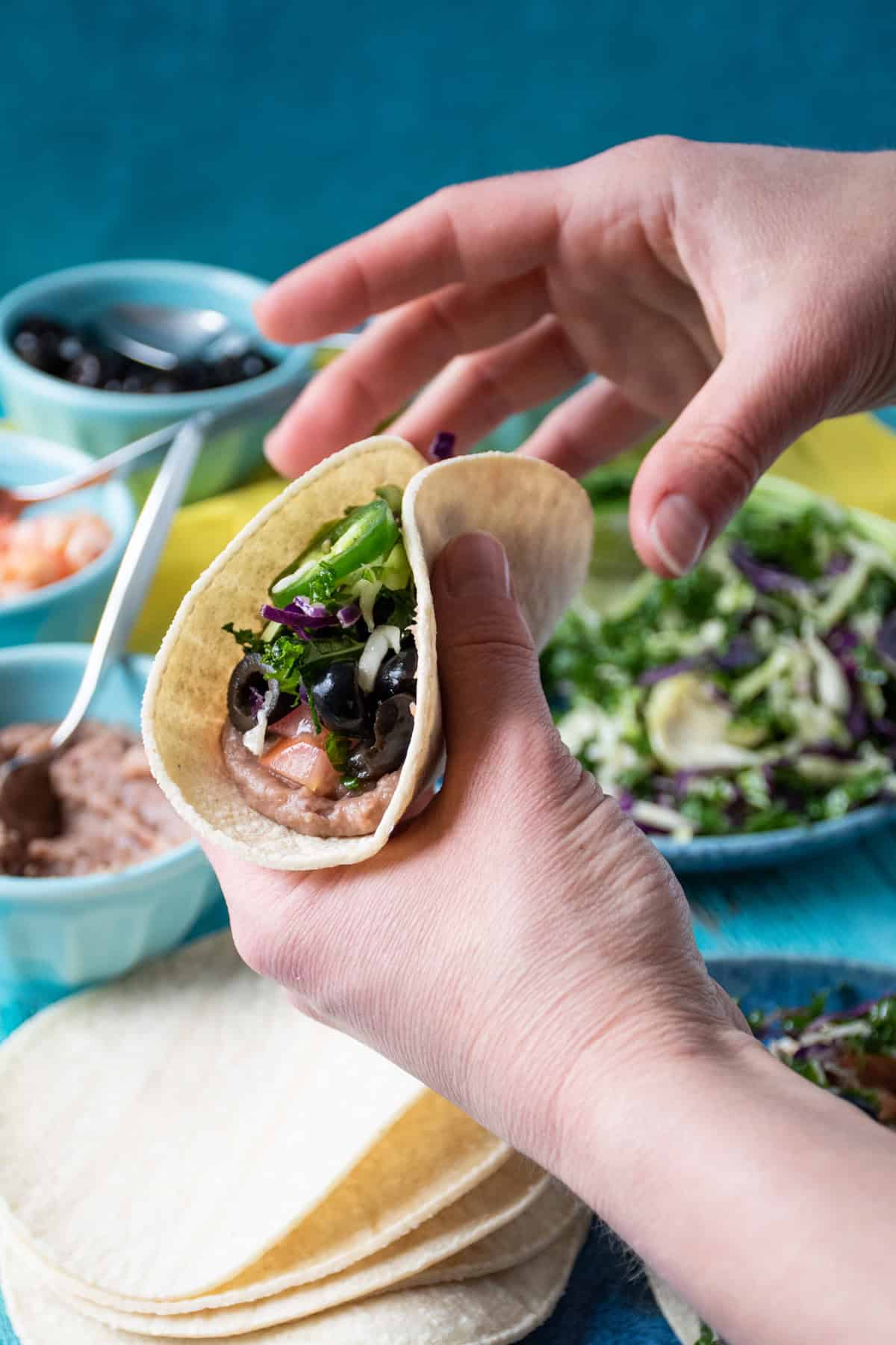 Two hands holding a taco, with all the ingredients to assemble visible in the background.
