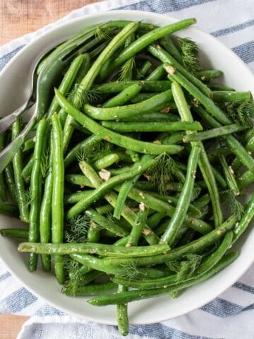 Roasted green beans in a bowl with serving utensils, garnished with fresh dill.