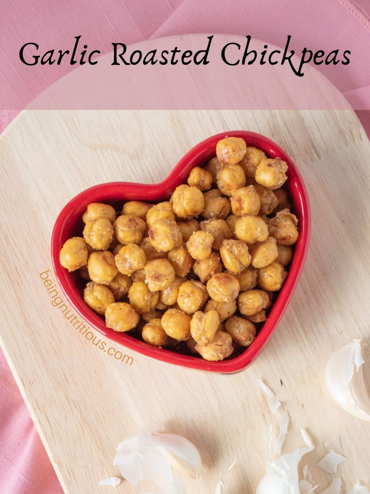 Crispy chickpeas in a heart shaped bowl, with raw garlic cloves around. Text overlay: Garlic Roasted Chickpeas.