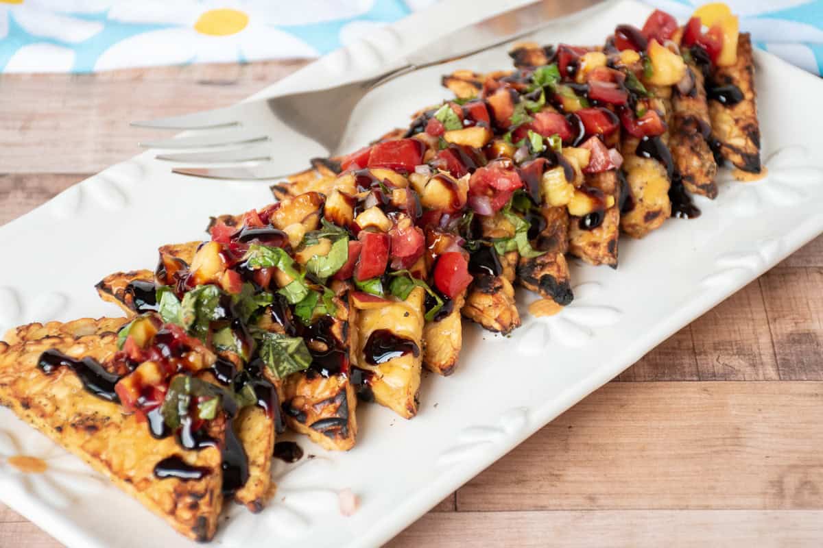 Grilled tempeh triangles in a line on a plate, topped with peach relish and drizzled with balsamic glaze.