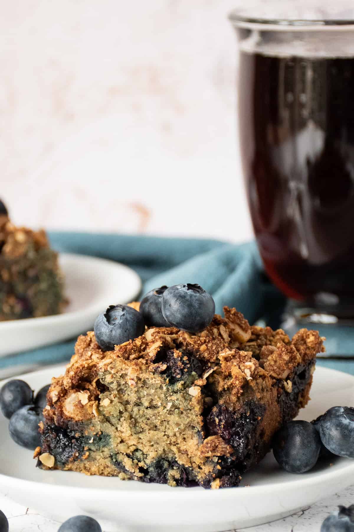 Slice of coffee cake on a plate, garnished with blueberries, with a cup of coffee in the background.