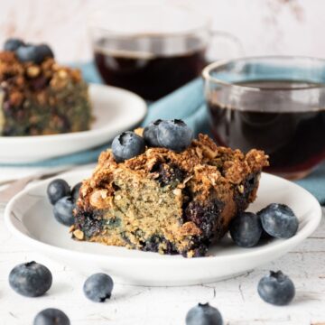 Slice of coffee cake on a plate, garnished with blueberries, with another slice of cake and two cups of coffee in the background.