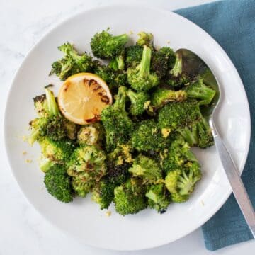 Grilled broccoli on a serving plate, garnished with a grilled lemon half, lemon zest and a stem of thyme.
