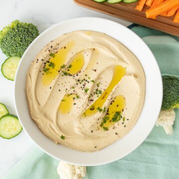 Bowl of creamy hummus, garnished with an olive oil drizzle, and a sprinkle of parsley, black sesame seeds, and sumac.