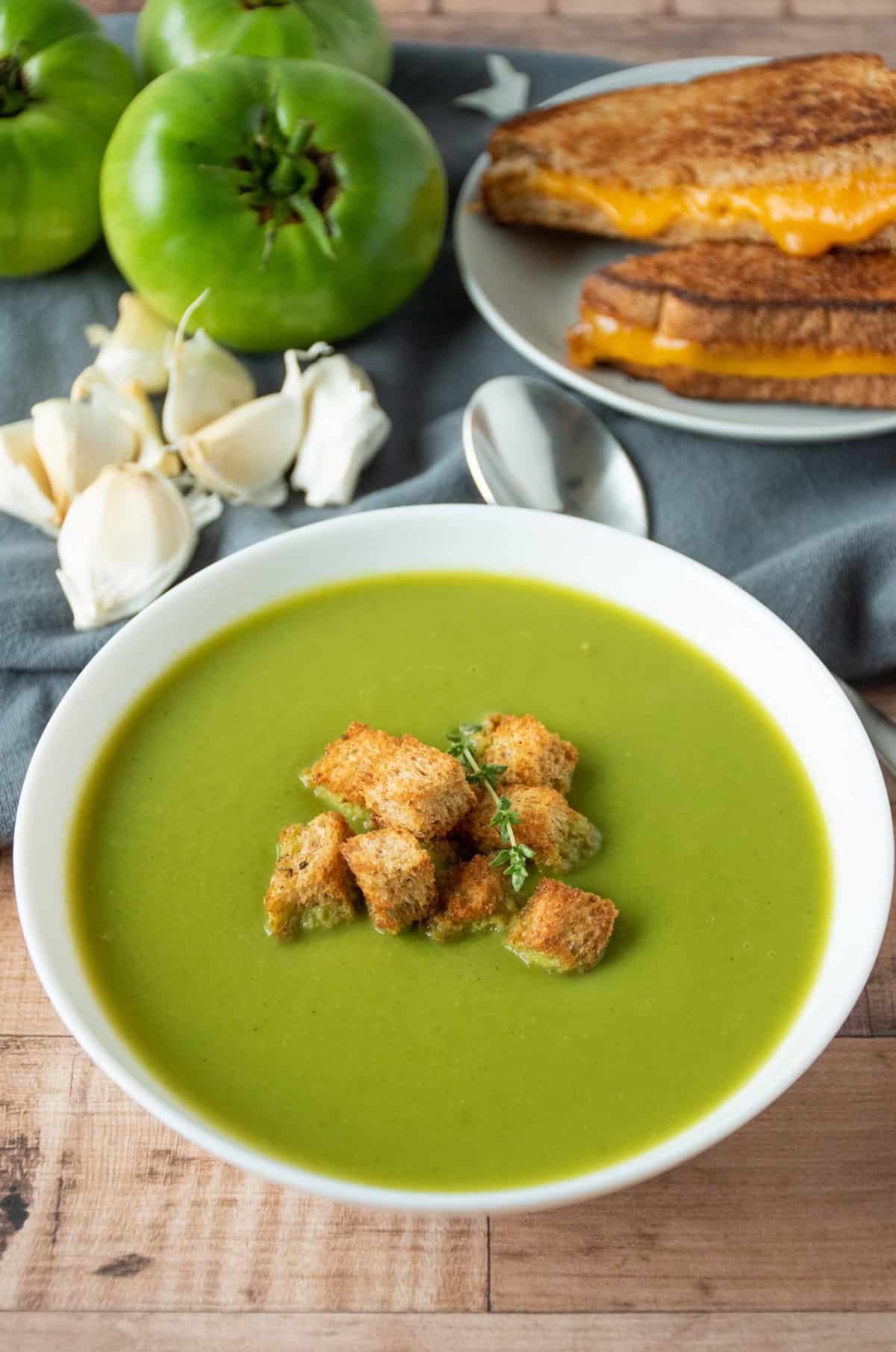 Green soup in a bowl, garnished with homemade croutons and fresh thyme. A grilled cheese sandwich and fresh garlic and green tomatoes are in the background.