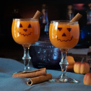 Two small wine glasses with jack-o-lantern face on them, with an orange beverage inside.