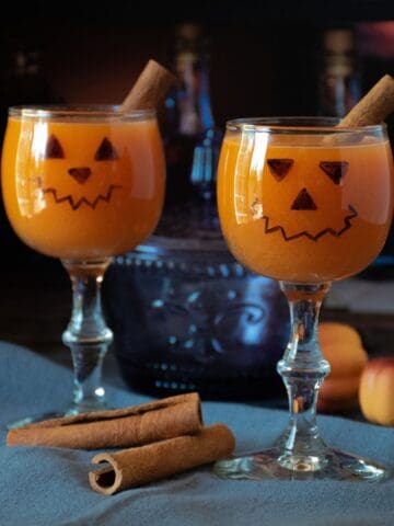 Two small wine glasses with jack-o-lantern face on them, with an orange beverage inside.