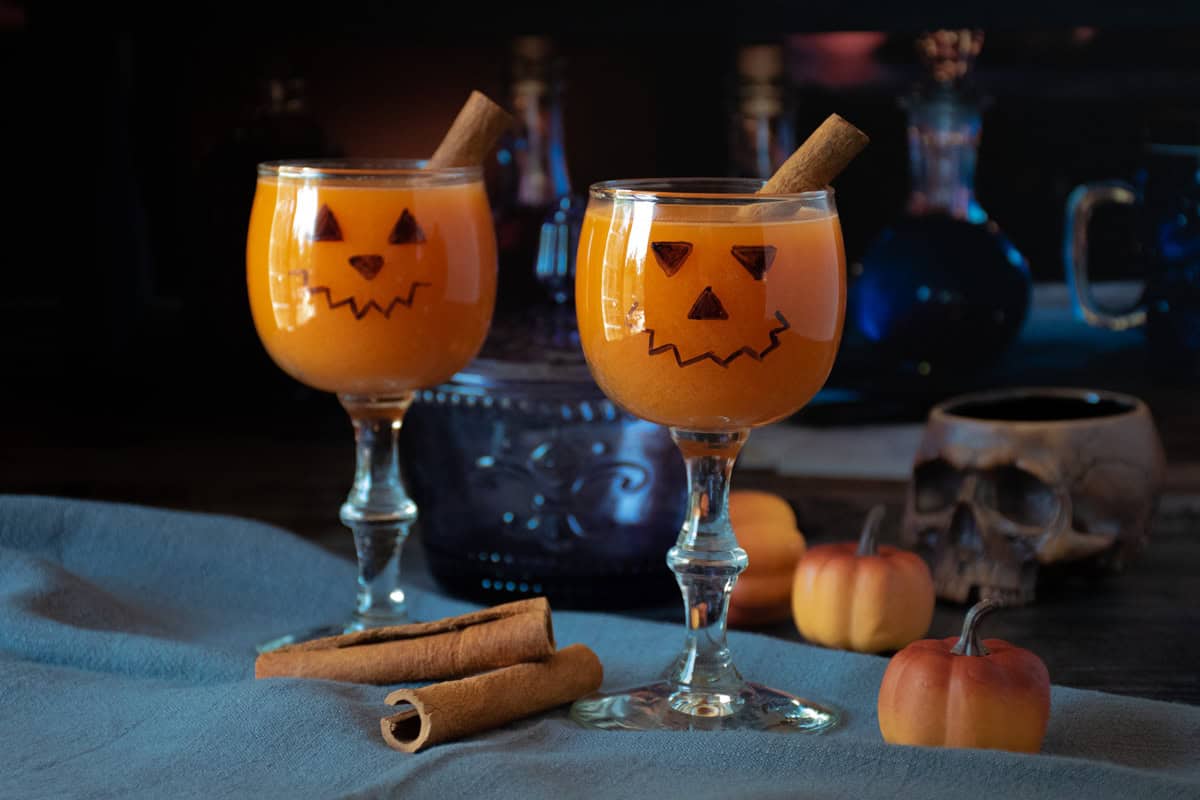 Two small wine glasses with jack-o-lantern face on them, with an orange beverage inside.