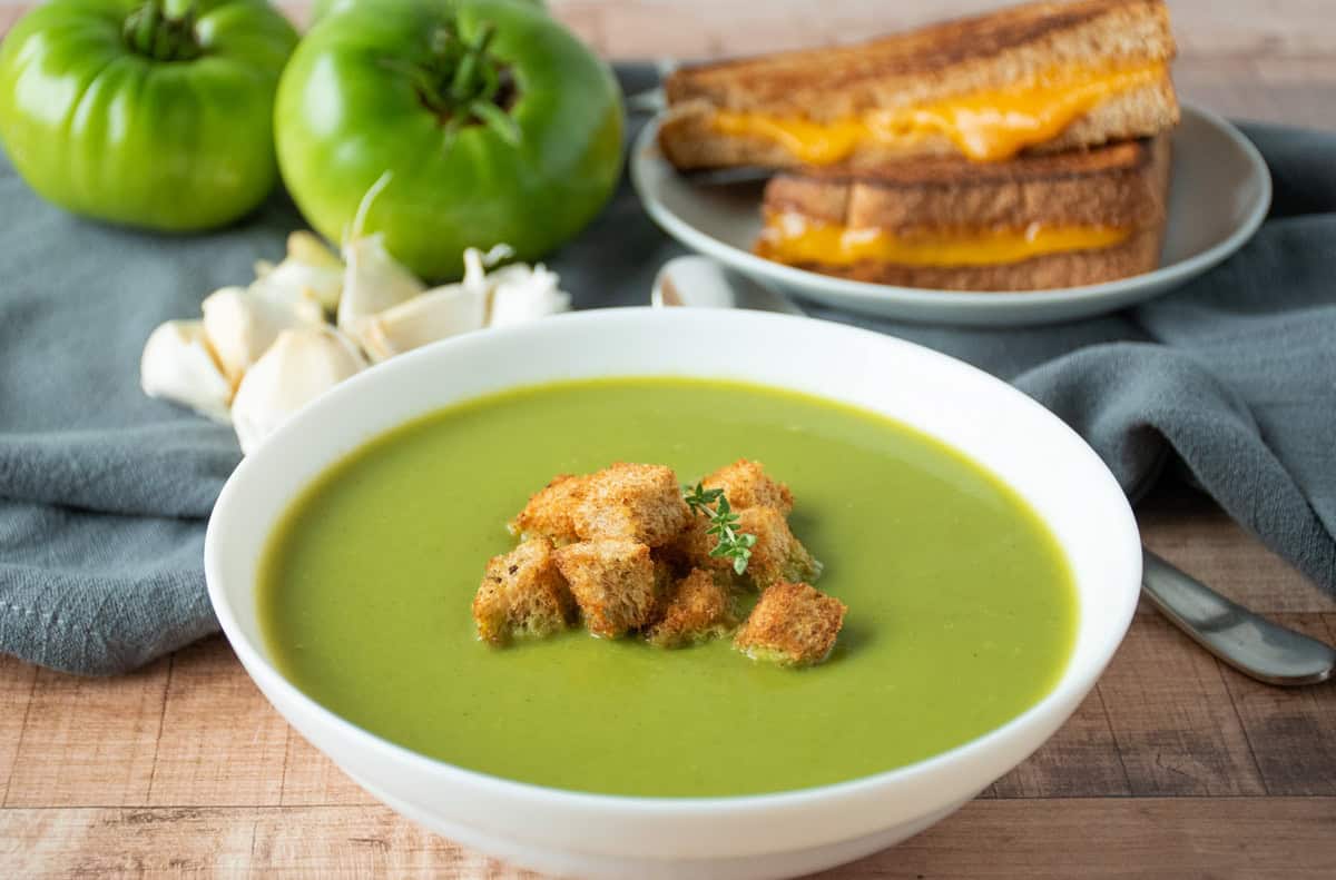 Green soup in a bowl, garnished with homemade croutons and fresh thyme. A grilled cheese sandwich and fresh garlic and green tomatoes are in the background.