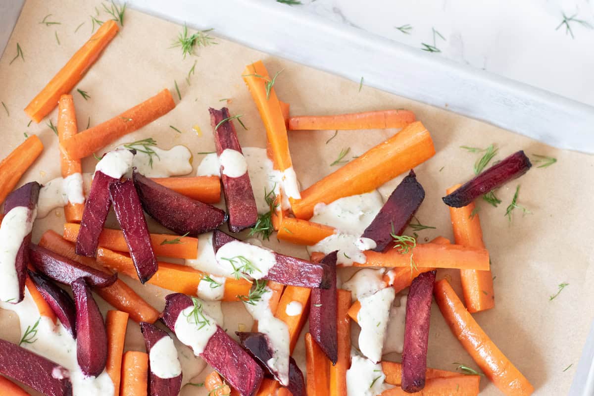 Roasted beets and carrots, on a baking sheet, cut into large matchsticks, drizzled with a white sauce and sprinkled with chopped dill.