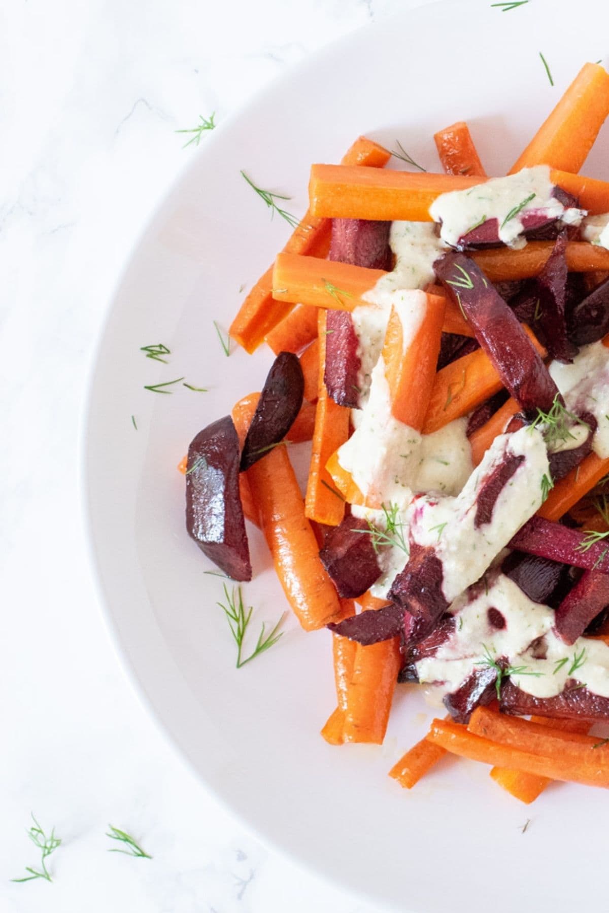 Roasted beets and carrots on a dish, cut into large matchsticks, drizzled with a white sauce and sprinkled with chopped dill.