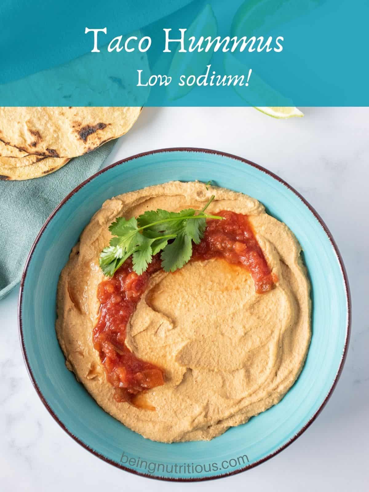 Bowl of hummus, garnished with salsa and cilantro. Cut limes and corn tortillas surround the bowl. Text overlay: Taco hummus; low sodium!