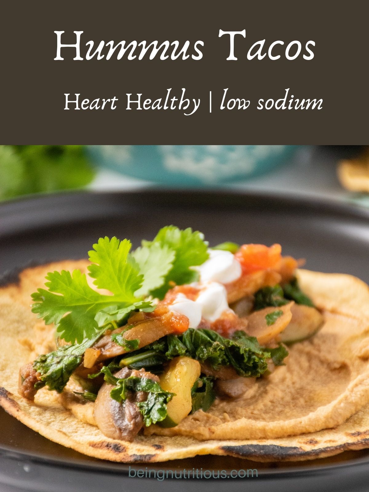Tortilla, flat on a plate, smeared with hummus and loaded with sauteed mushrooms, zucchini, and kale, garnished with cilantro. Text overlay: Hummus Tacos; heart healthy, low sodium.