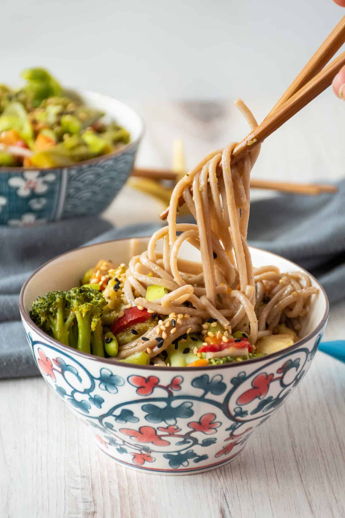 Bowl of noodles and vegetables, with some noodles being lifted from the bowl with chopsticks.