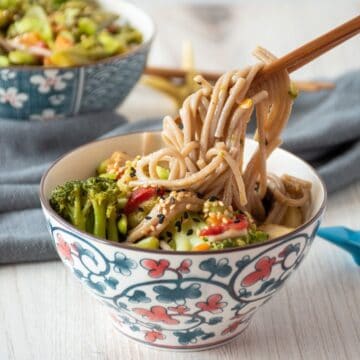 Bowl of noodles and vegetables, with some noodles being lifted from the bowl with chopsticks.