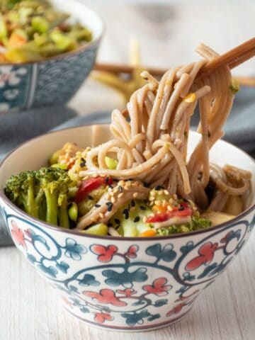 Bowl of noodles and vegetables, with some noodles being lifted from the bowl with chopsticks.