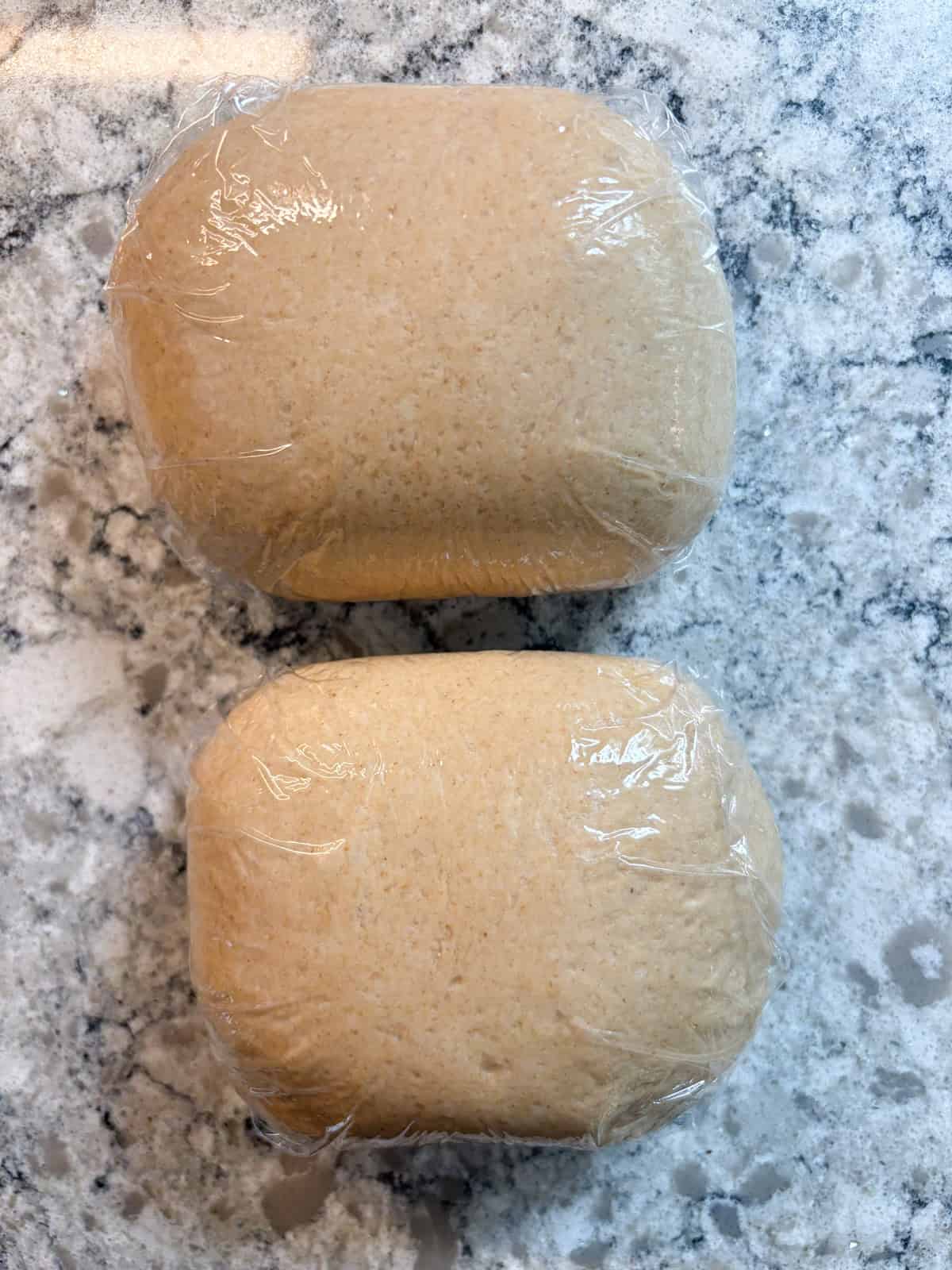 Two balls of raw pizza dough wrapped in plastic wrap.