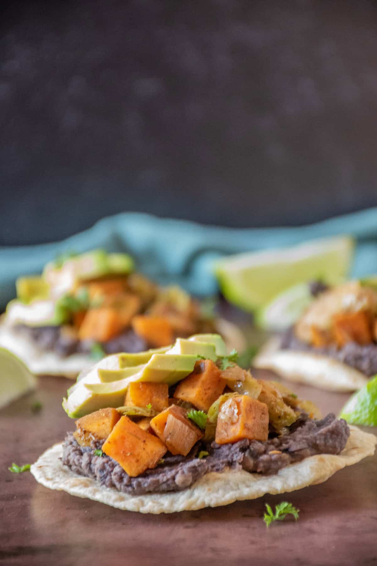 Side image of a tostada, with black bean puree and sautéed sweet potatoes and Brussels sprouts, garnished with avocado and minced parsley.
