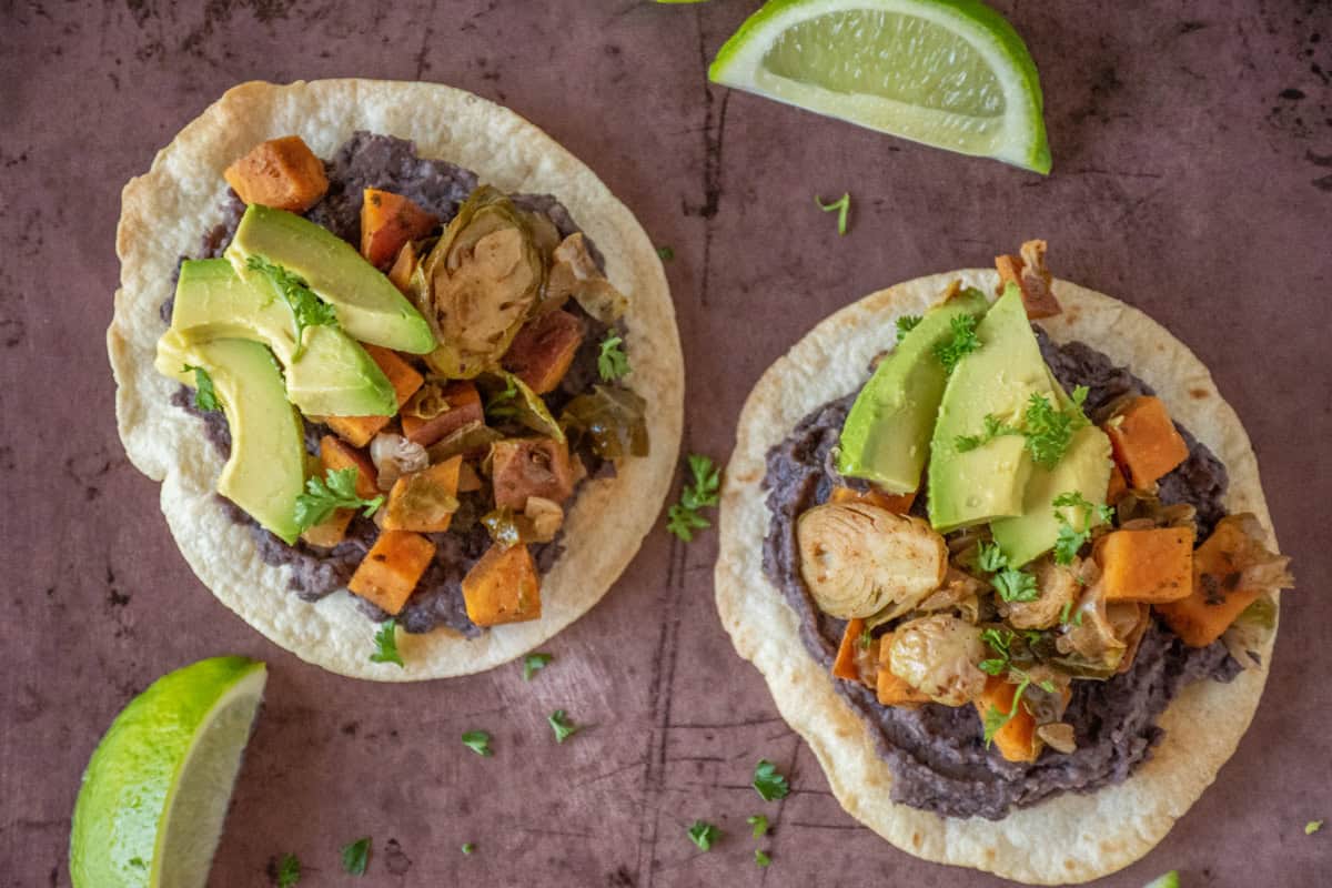 Overhead image of 2 tostadas, with black bean puree and sautéed sweet potatoes and Brussels sprouts, garnished with avocado and minced parsley.