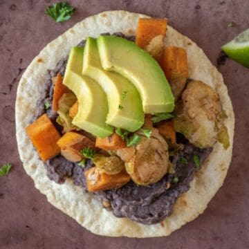 Overhead image of a tostada, with black bean puree and sautéed sweet potatoes and Brussels sprouts, garnished with avocado.