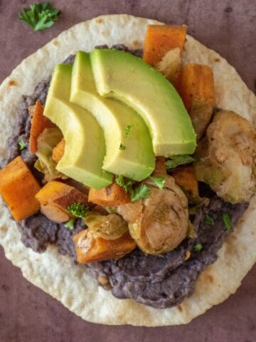 Overhead image of a tostada, with black bean puree and sautéed sweet potatoes and Brussels sprouts, garnished with avocado.