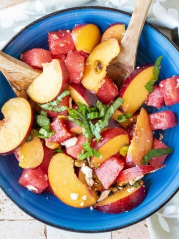 Overhead image of fruit salad with wooden serving utensils, in a blue bowl.