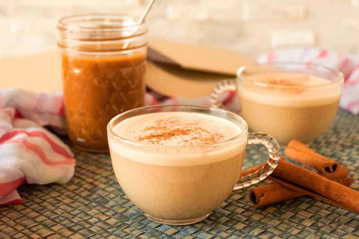 A glass mug containing a pumpkin spice latte made with healthy pumpkin spice syrup, seen in the background, along with another latte drink.