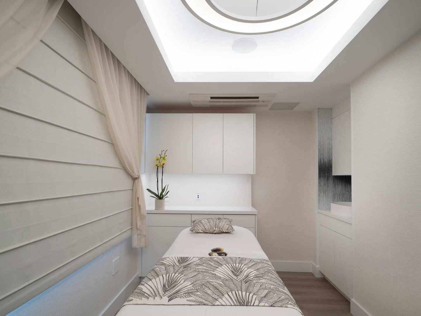 Narrow spa treatment room with white wall panels and an illuminated ceiling recess.