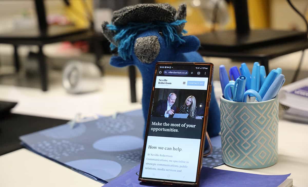 Dundee office desk with plush bear, smartphone displaying Neville Robertson Communications website,.