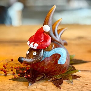 Bernsteinfarbenes Weihnachtsobjekt Igel SANTA SPIKE mit roter Weihnachtsmütze auf einem mit Laub und roten Beeren dekorierten Holztisch.