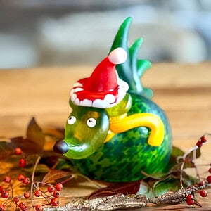 Grünes Weihnachtsobjekt Igel SANTA SPIKE mit roter Weihnachtsmütze auf einem mit Laub und roten Beeren dekorierten Holztisch.
