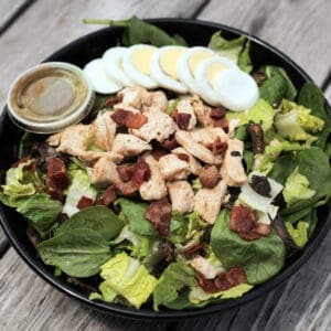 Fresh salad with chicken, bacon, boiled eggs, and mixed greens in a black bowl.