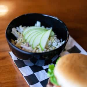 Fresh coleslaw with sliced green apple in a black bowl on checkered paper.