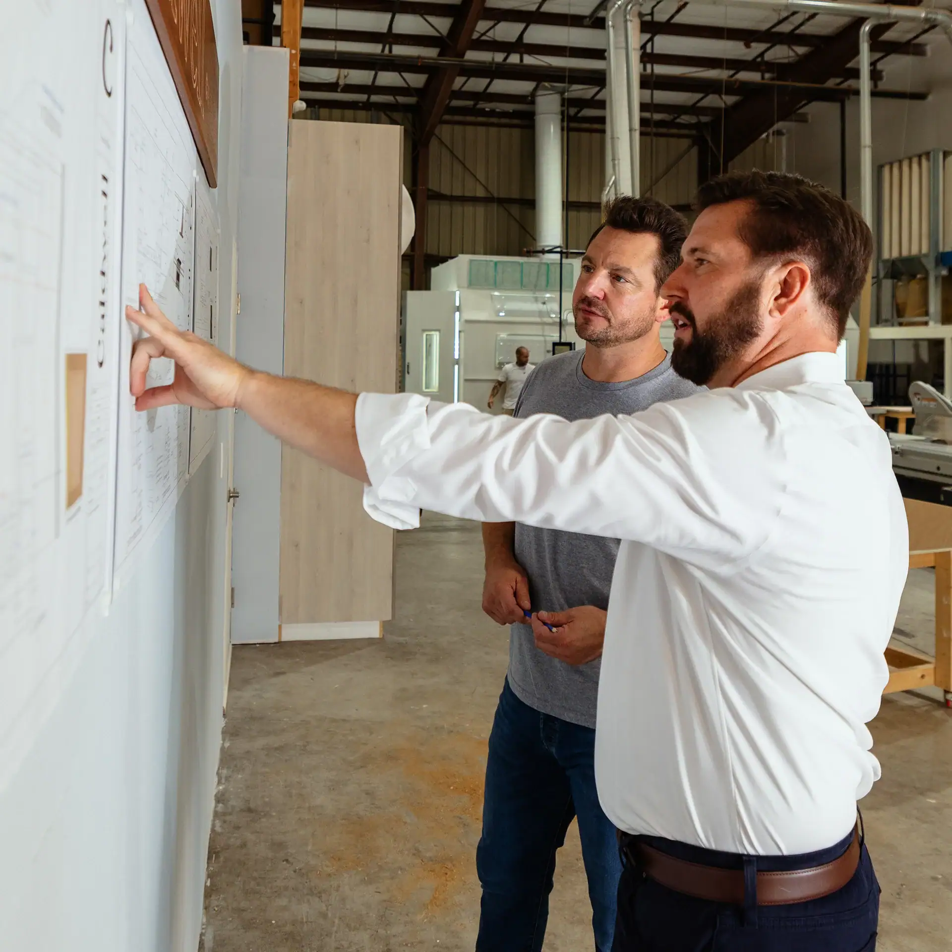 Two men in business attire standing before a whiteboard, discussing or explaining blueprints placed on wall.