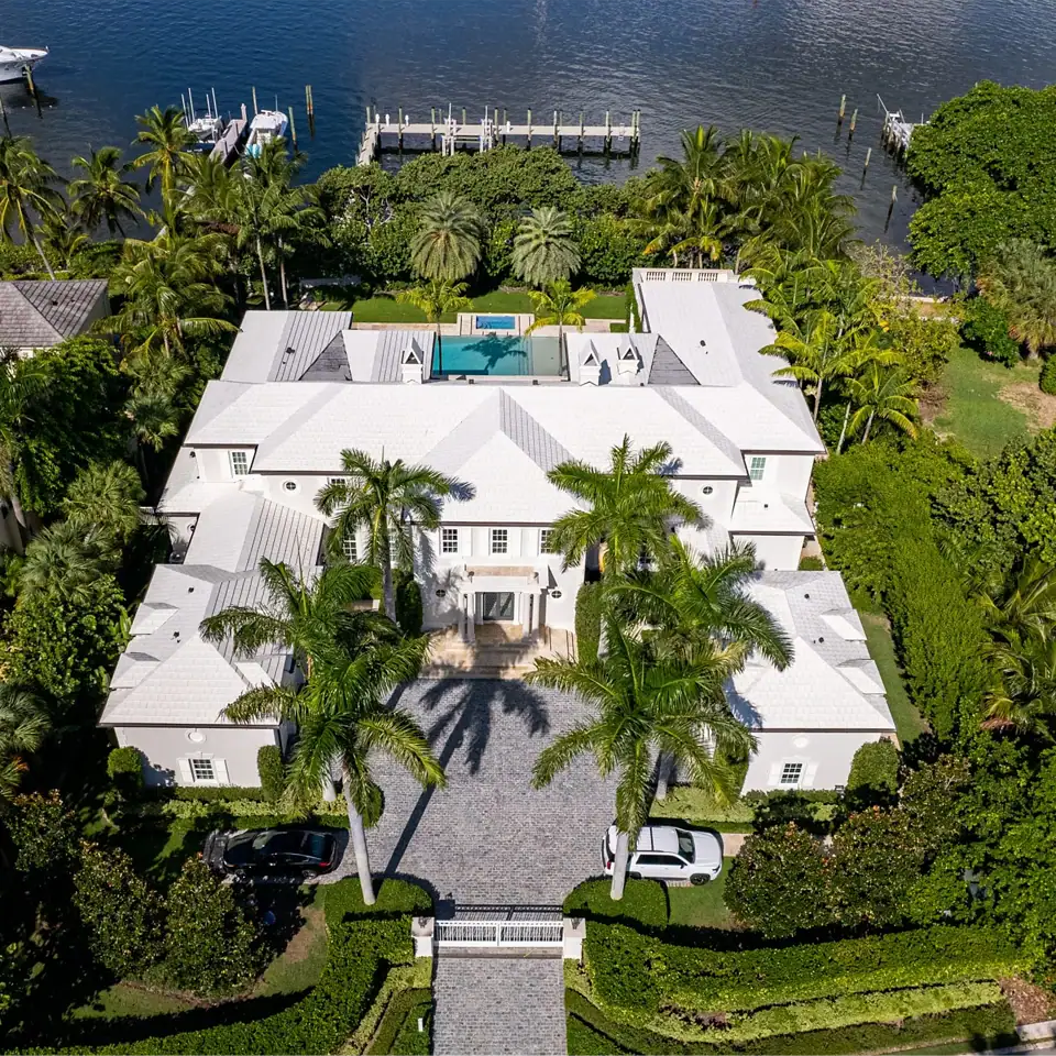 Aerial view of an expansive Bermuda Colonial style home surrounded by lush palm trees and a serene body of water.