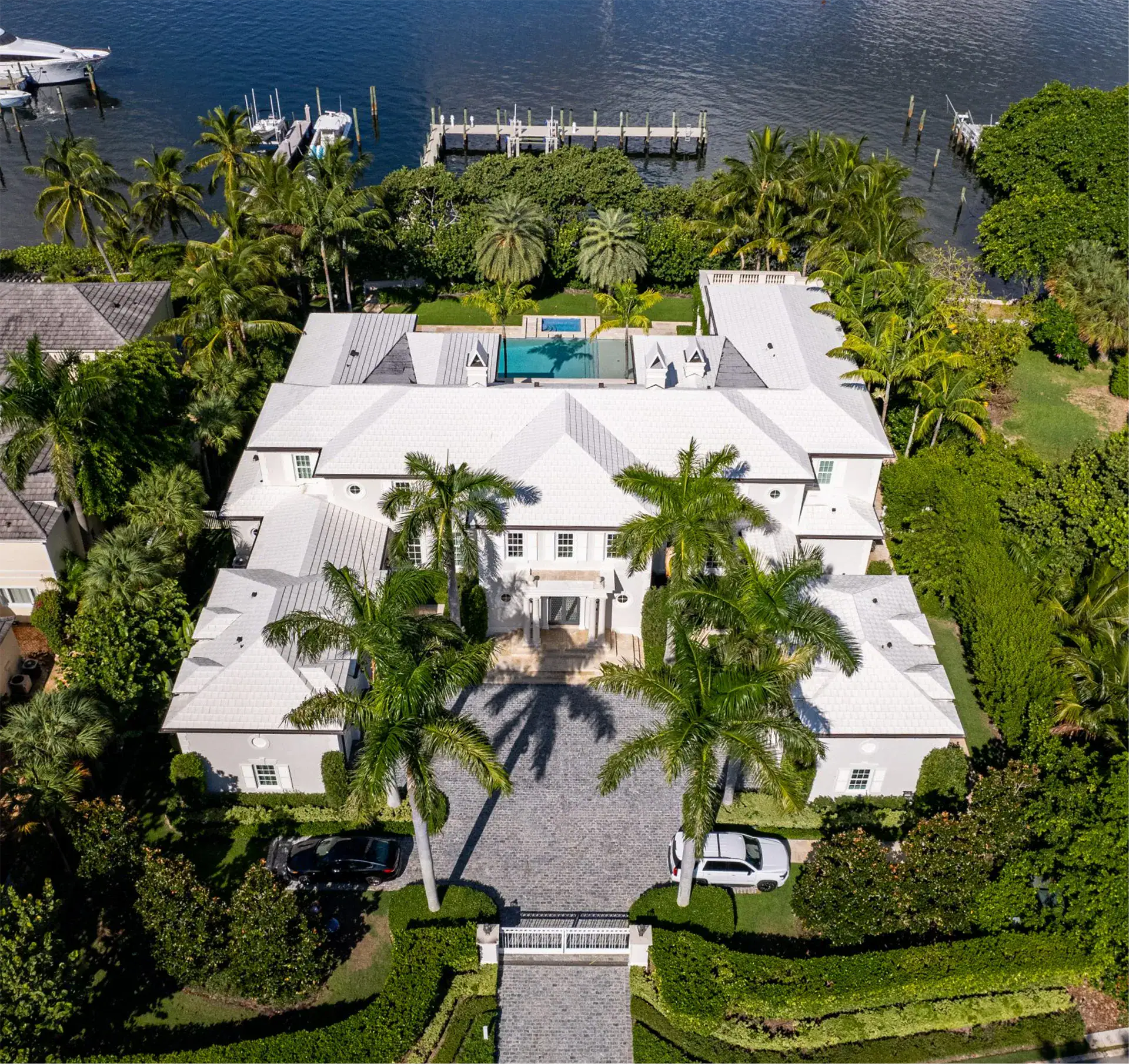 Aerial view of an expansive Bermuda Colonial style home surrounded by lush palm trees and a serene body of water.