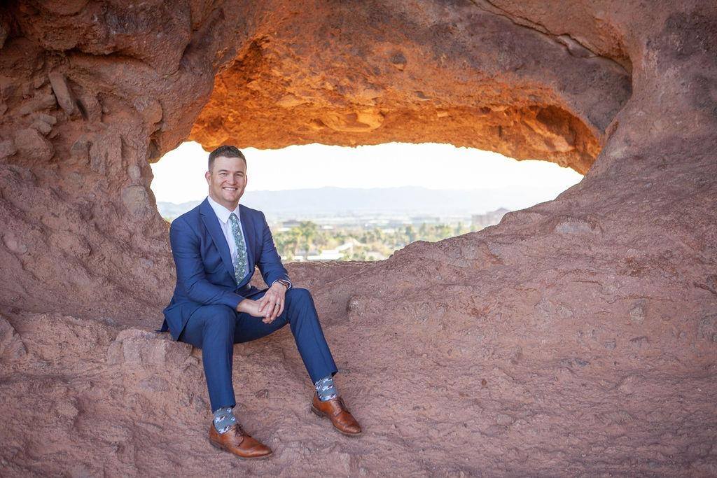 A man in a blue suit sitting inside a natural rock formation with a scenic background.