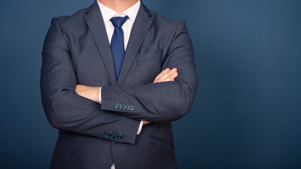 A person in a navy suit with crossed arms against a dark blue background.