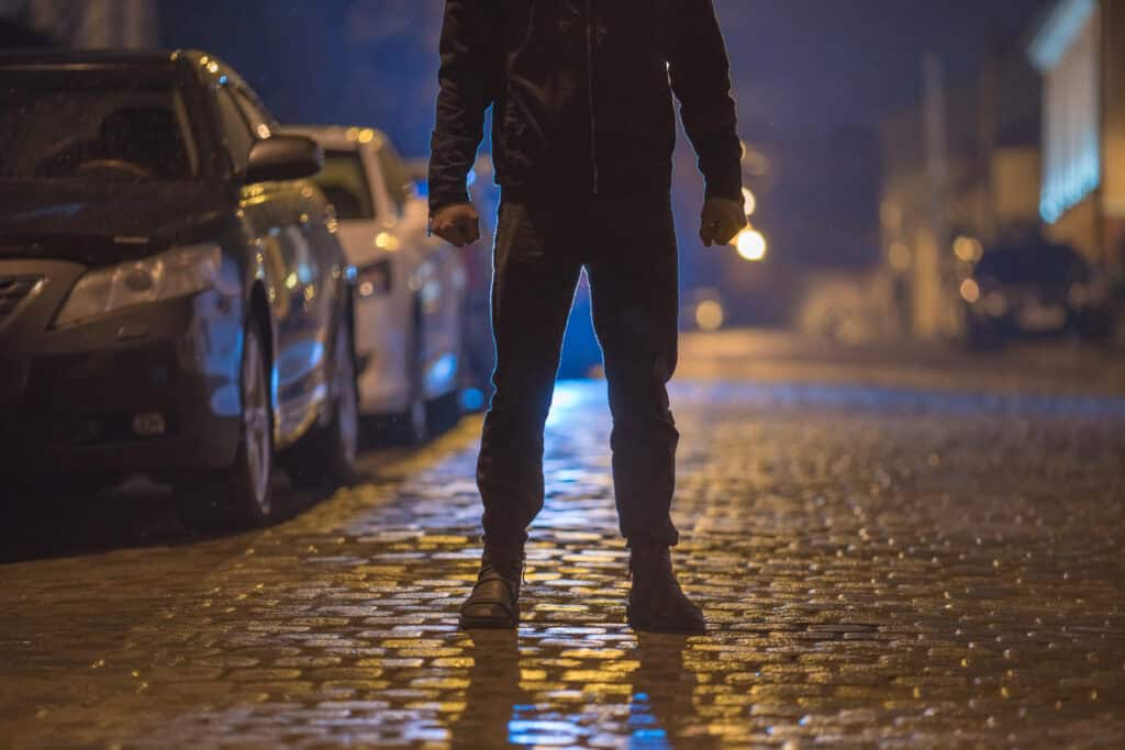 A person standing on a cobblestone street at night with parked cars nearby.