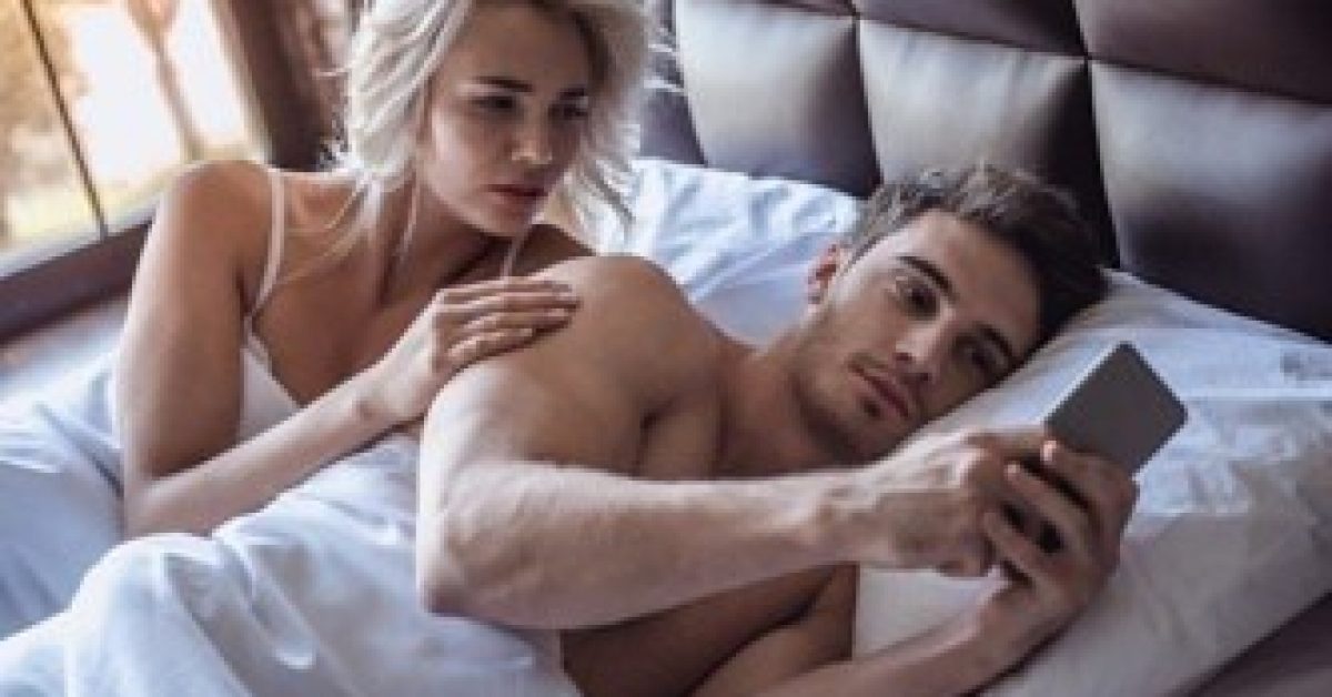 Beautiful young couple is lying in bed, guy is using a smart phone, girl is looking with reproach