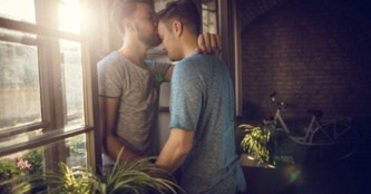 Loving gay men standing by the window while one of them is kissing his boyfriend in the forehead.